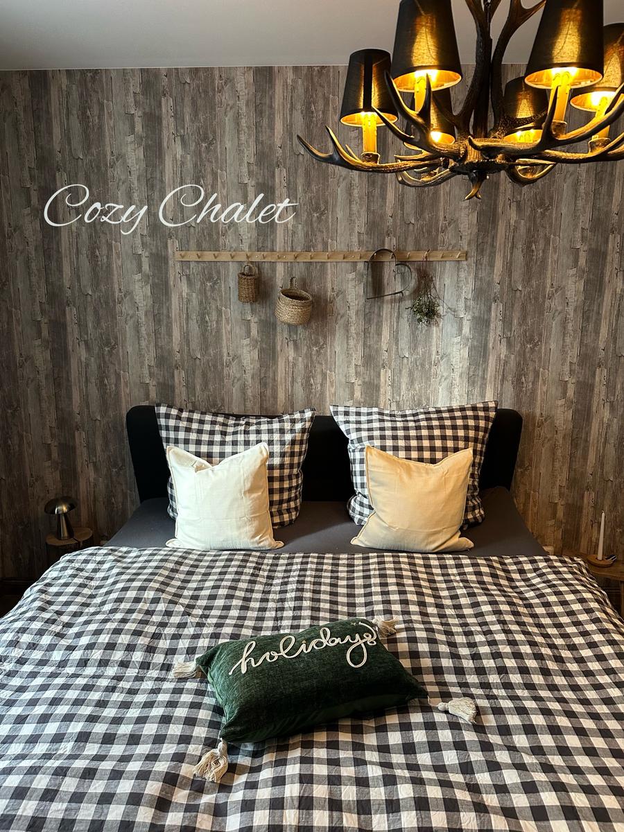 Schlafzimmer mit Holzwand, Bett mit kariertem Bettzeug und Hirschgeweih-Leuchter.
