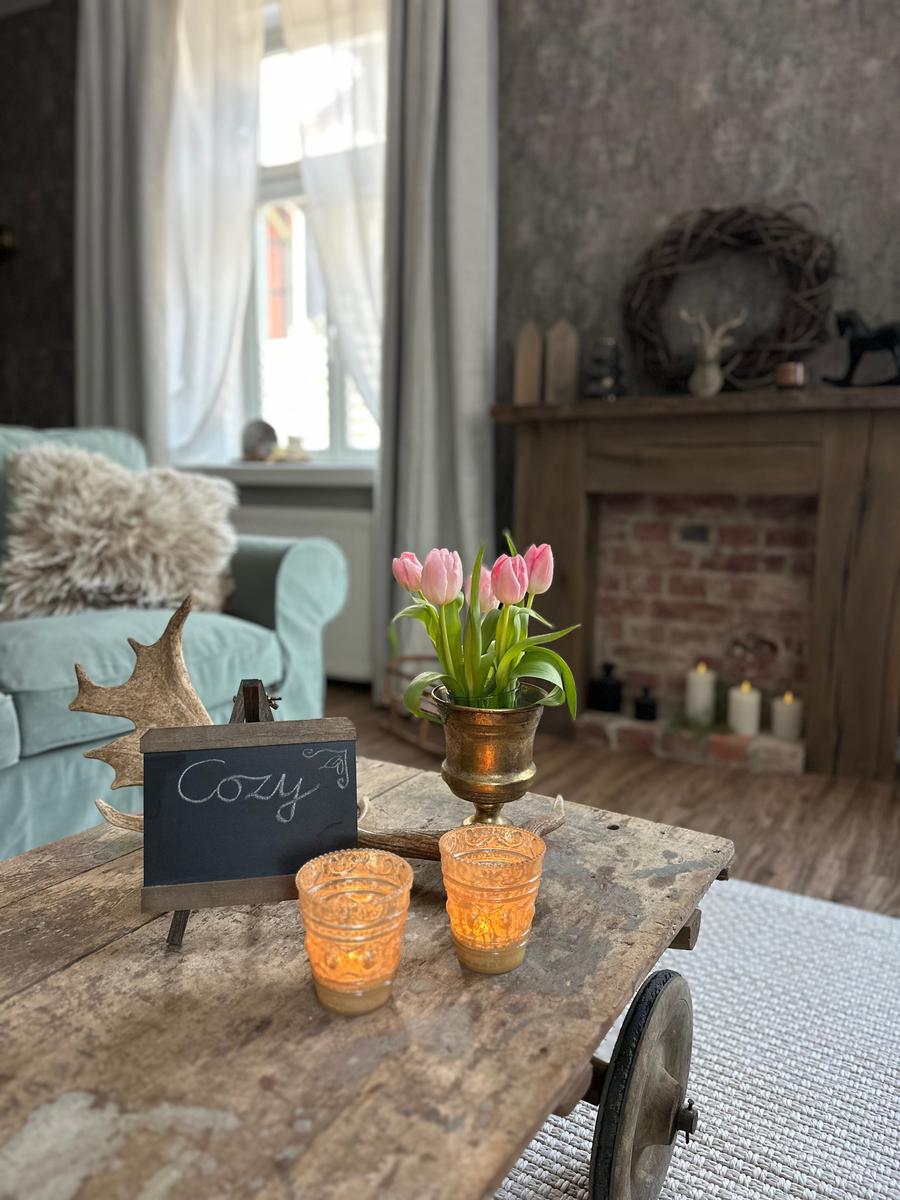 Holztisch mit Tulpen, Kerzen und Schild. Im Hintergrund Sofa und Kamin.