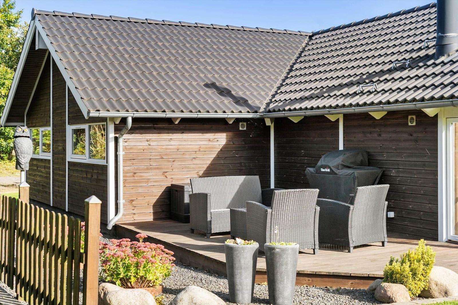 Holzhaus mit Terrasse, Gartenmöbeln und Grill. Umgeben von Pflanzen und Steinen.