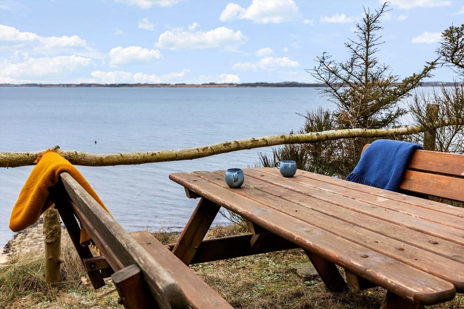 Holztisch und Bänke am Wasser mit Decken und Tassen.