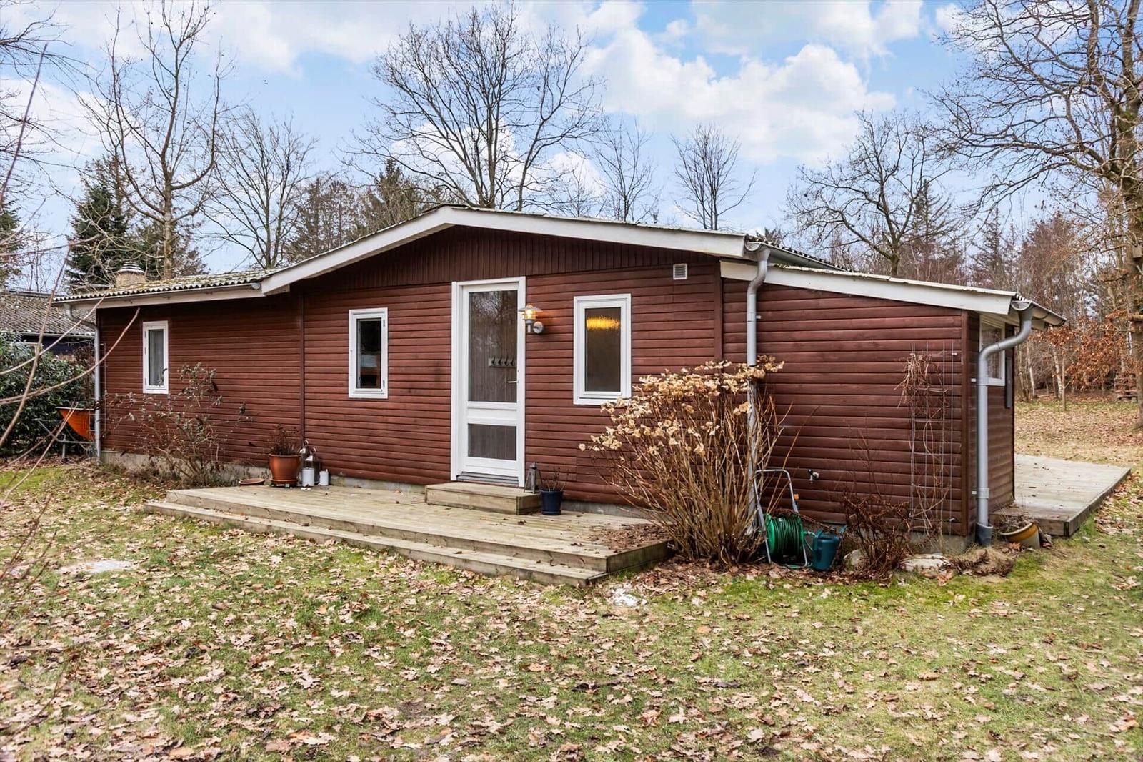 Ein kleines Holzhaus mit weißer Tür und Fenstern. Vorgarten mit Laub und Sträuchern.