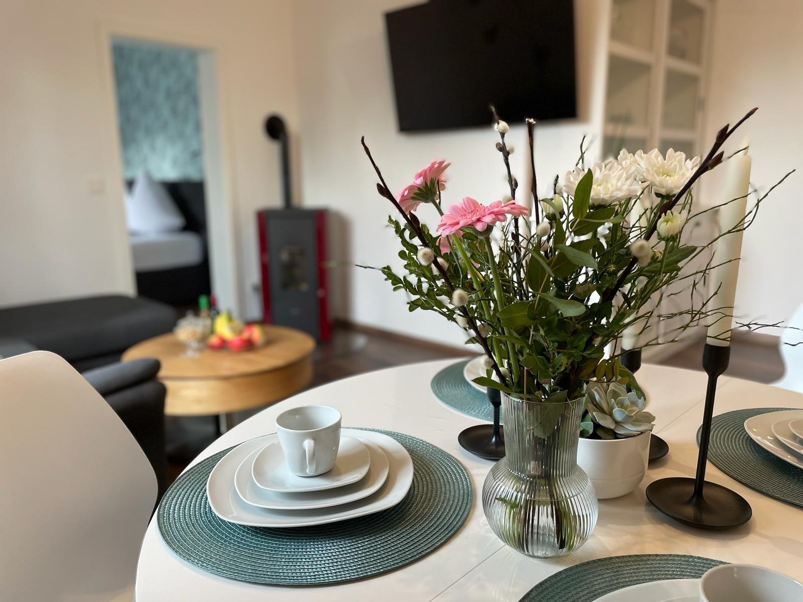 Esstisch mit Blumen, Tassen und Platten. Im Hintergrund Wohnzimmer mit Sofa und Kaminofen.