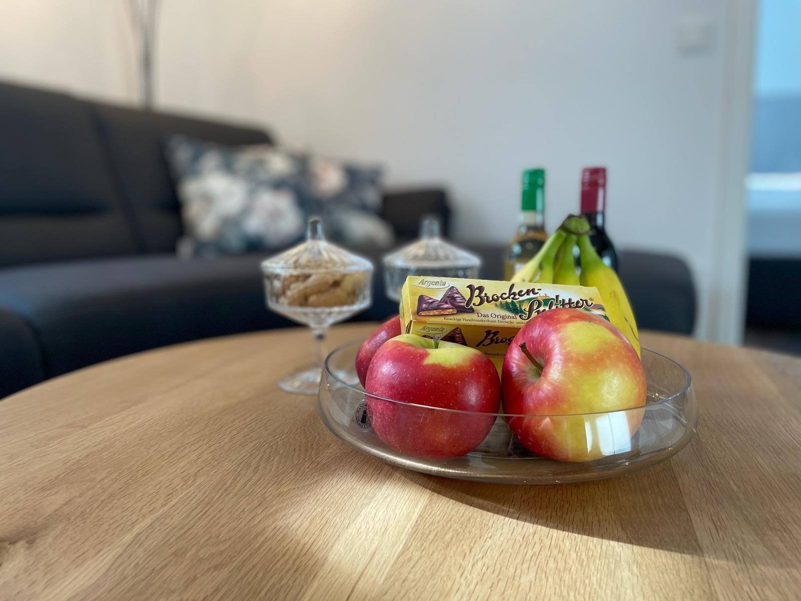 Auf dem Tisch stehen Äpfel, Bananen und Schokolade. Im Hintergrund ist ein Sofa.