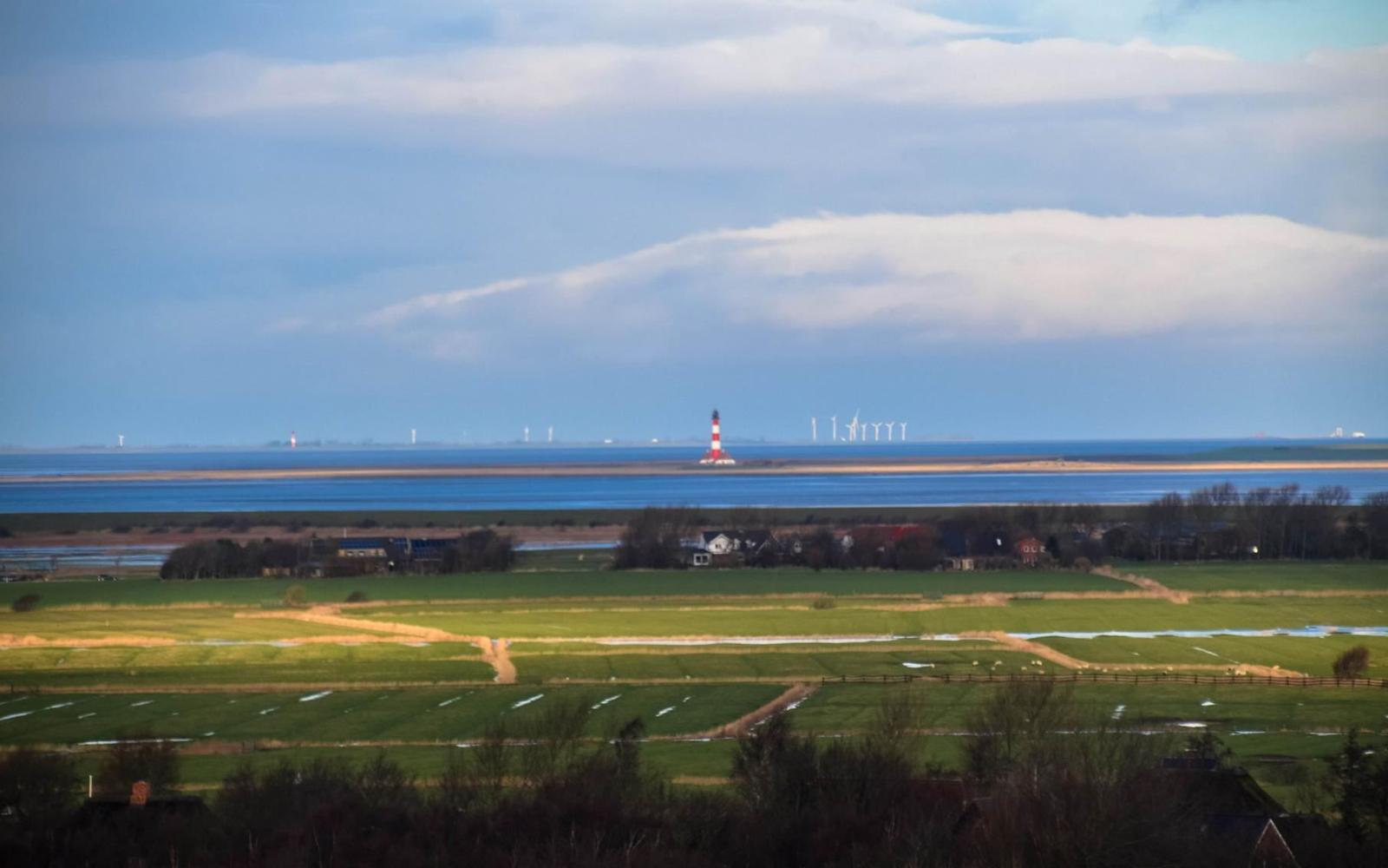 Weitläufige Landschaft mit Leuchtturm, Windkraftanlagen und grünen Feldern am Wasser.