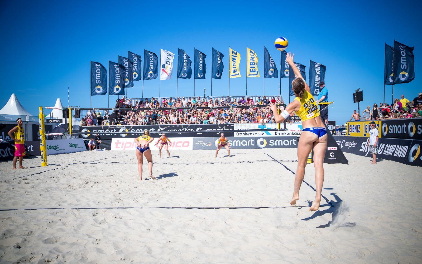 Beachvolleyball-Spiel mit Zuschauern und Sponsorenflaggen am Strand.