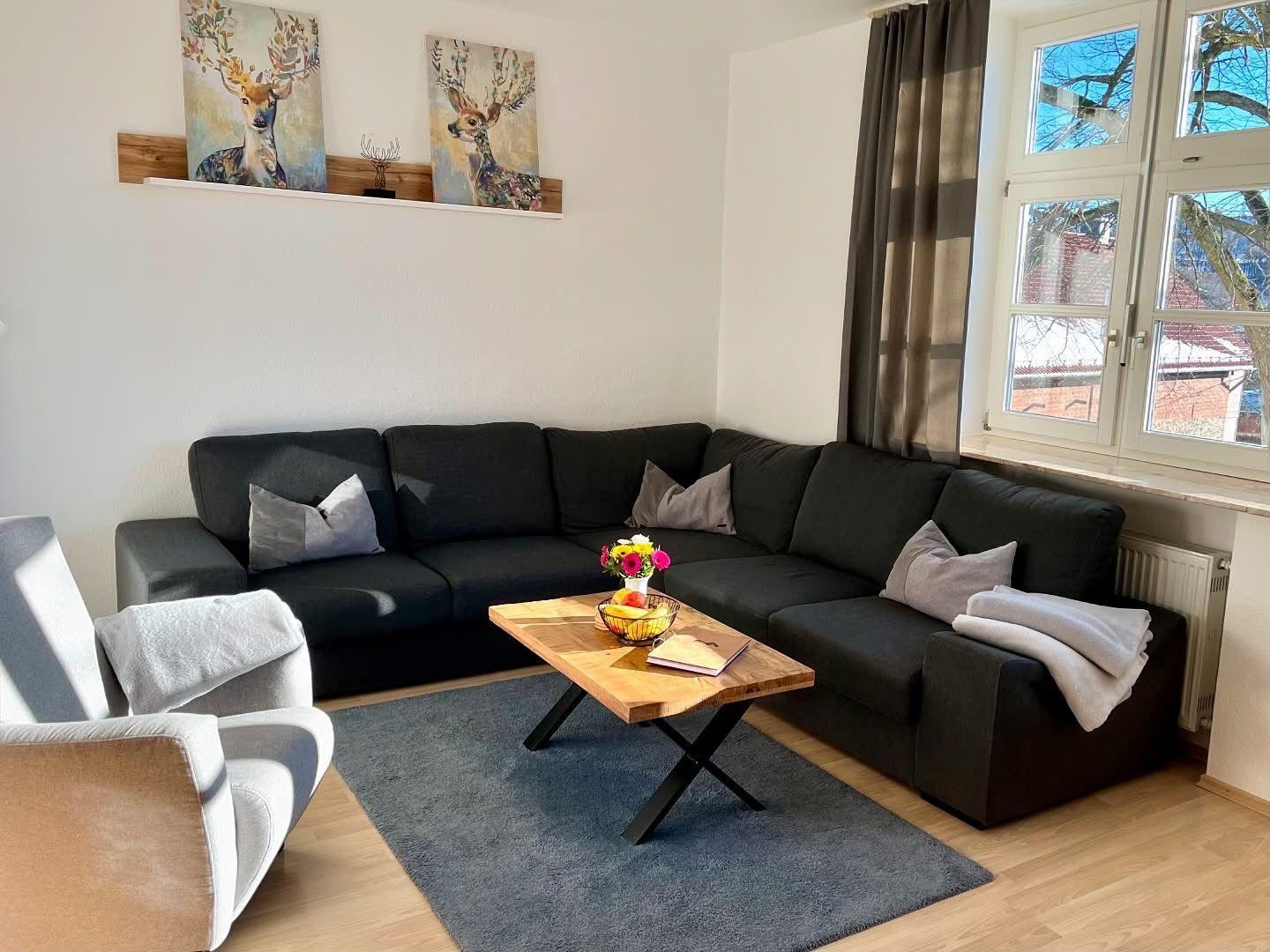 Ein Wohnzimmer mit schwarzer Couch, Holztisch und Fensterblick.