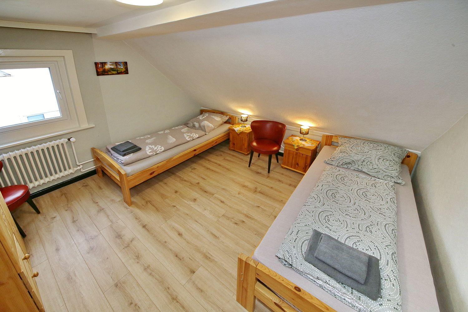 Zimmer mit zwei Betten, Holzboden und roter Sitzgelegenheit.