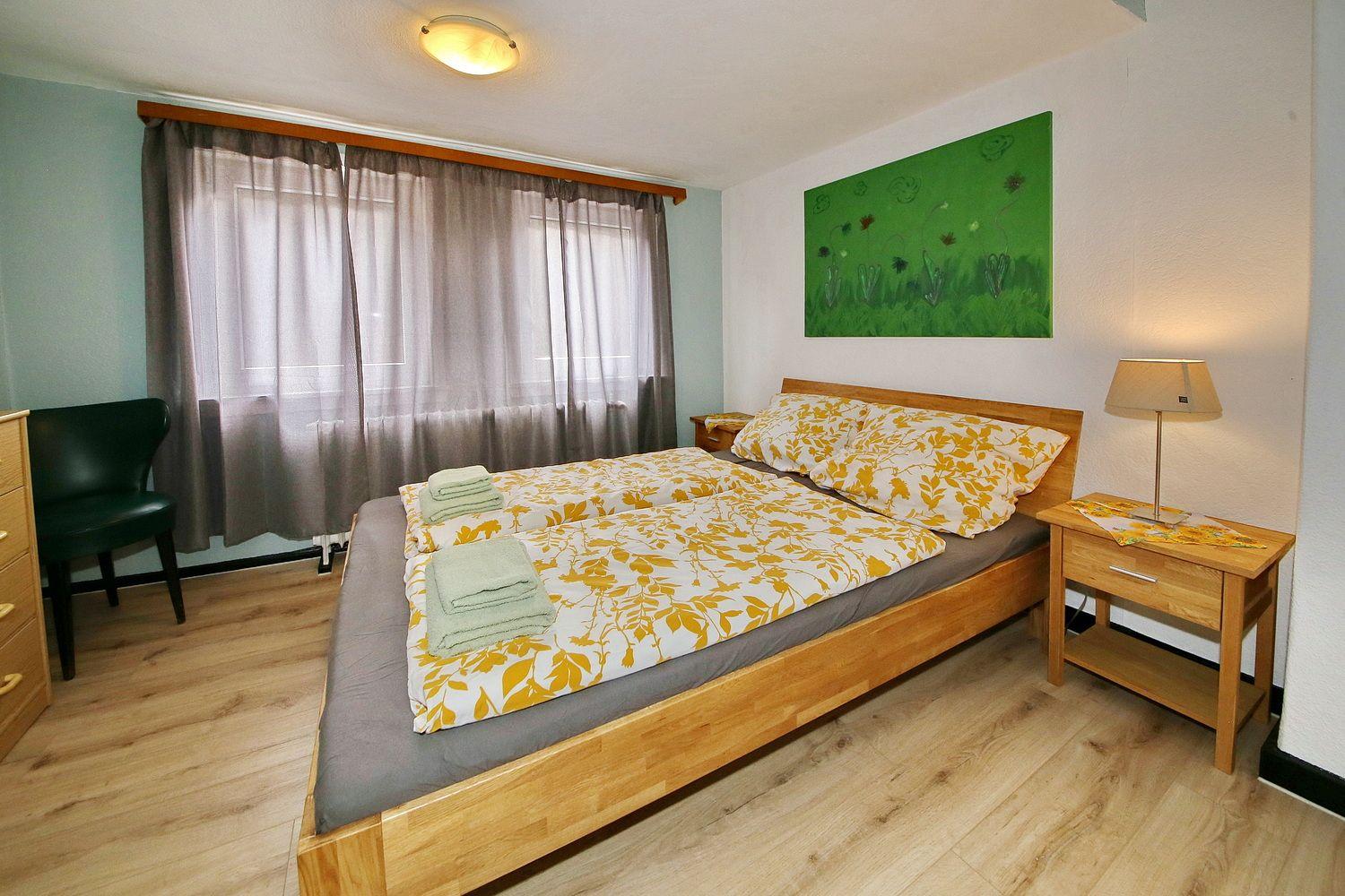 Schlafzimmer mit Holzbett, Nachttisch, Lampe und Fenster mit Vorhängen.