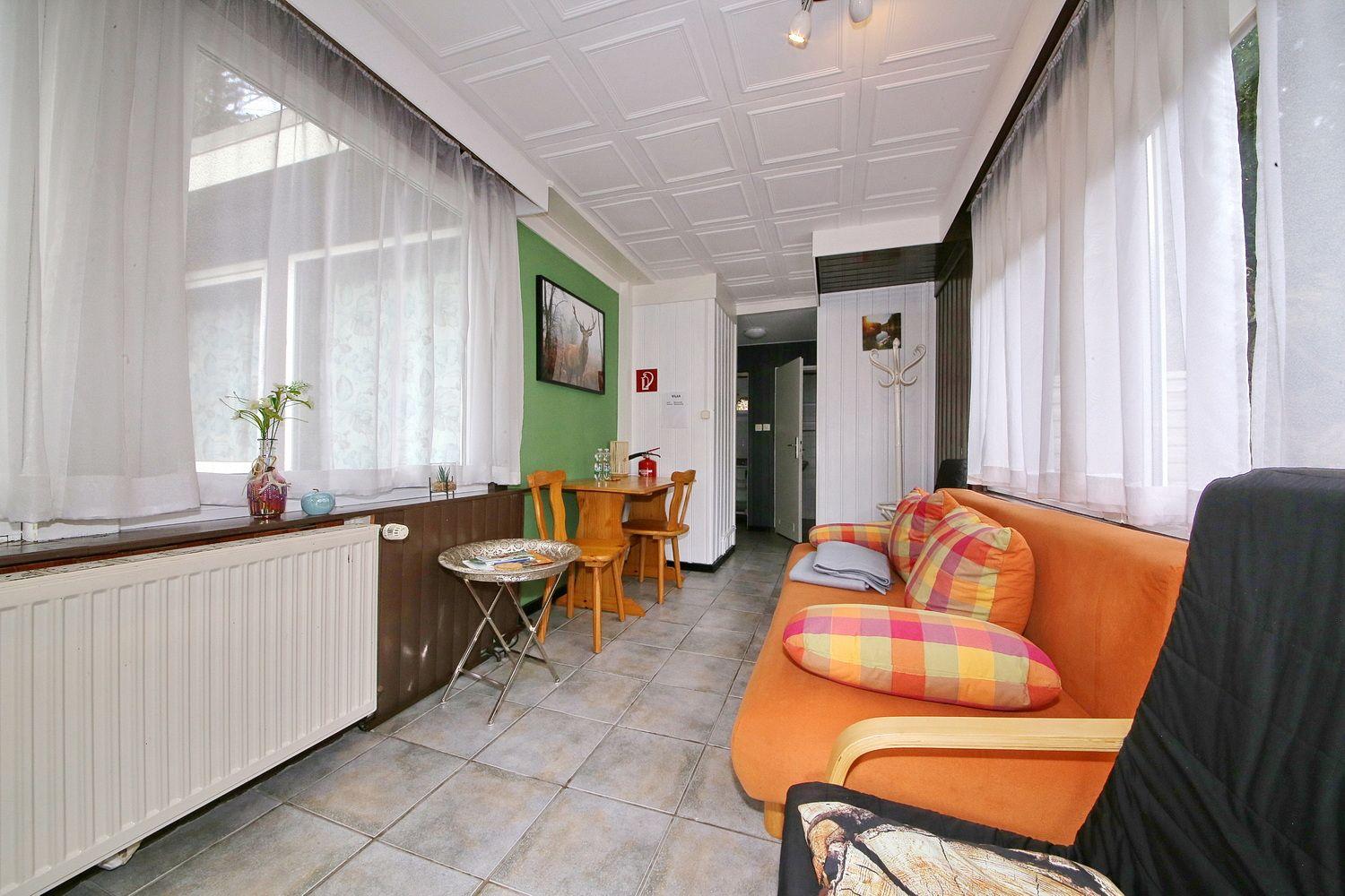 Wohnzimmer mit orangefarbenem Sofa, Tisch und Stühlen, Fenster mit Vorhängen und Kachelboden.