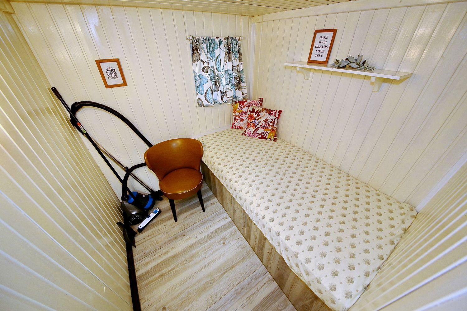 Ein Raum mit Bett, Stuhl und Fenster. Wandregal und Staubsauger sind sichtbar.