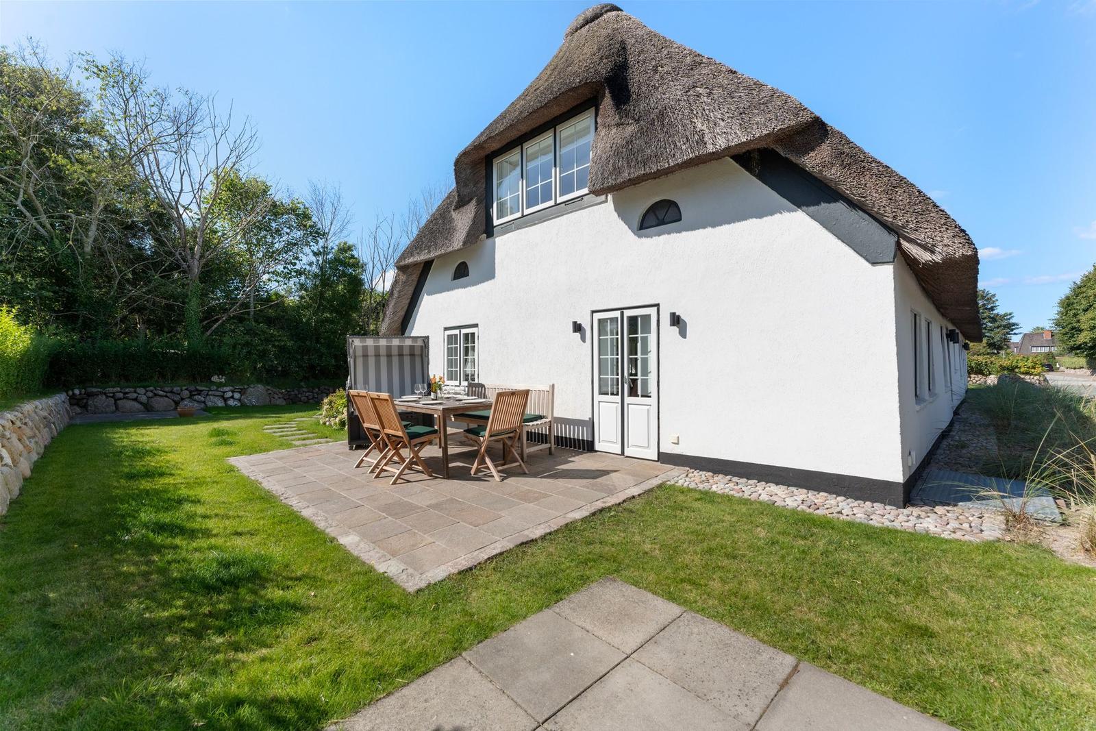 Haus mit Strohdach, Terrasse und Garten. Holztisch mit Stühlen.