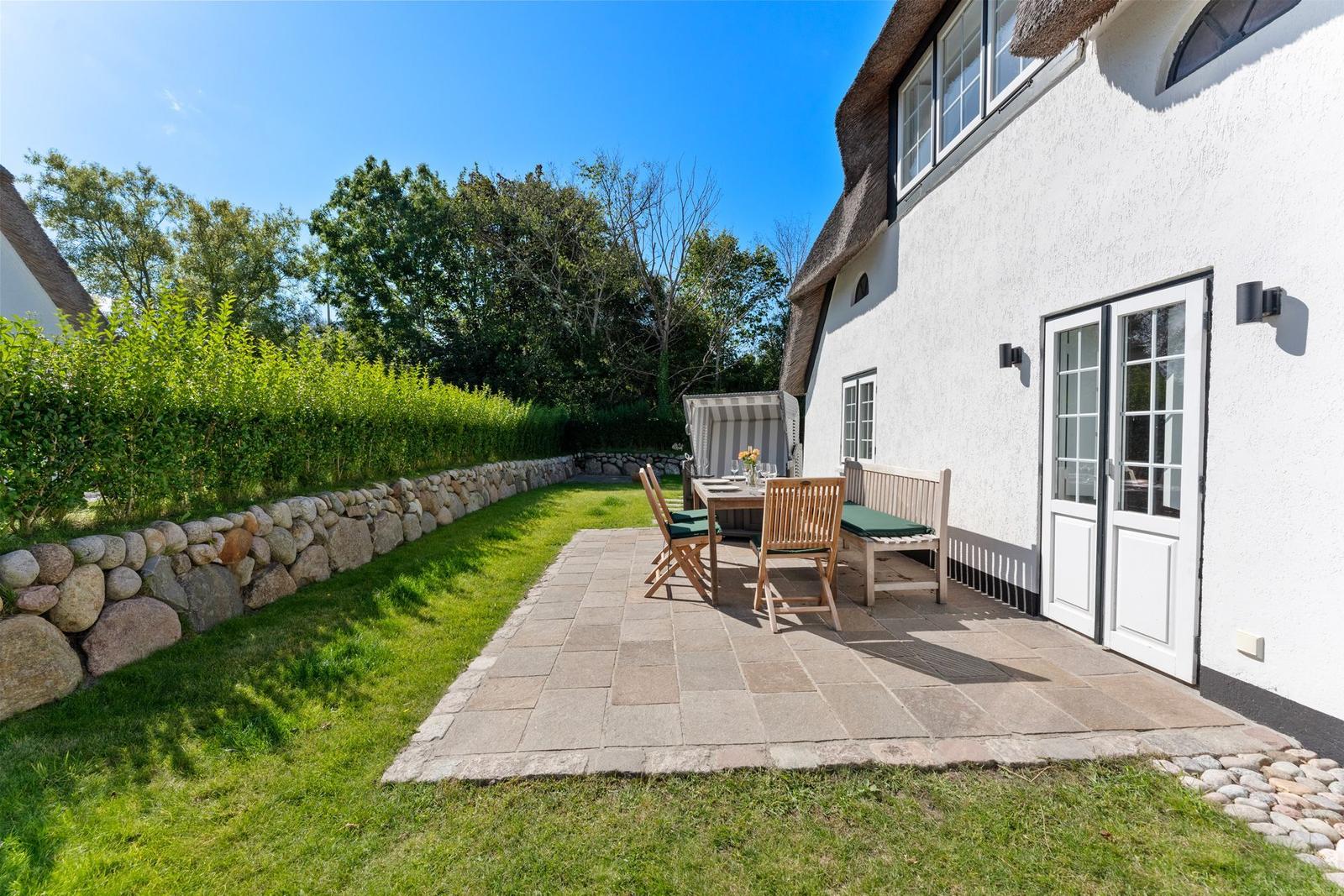 Terrasse mit Tisch, Stühlen und Bänken neben weißem Haus mit Strohdach.