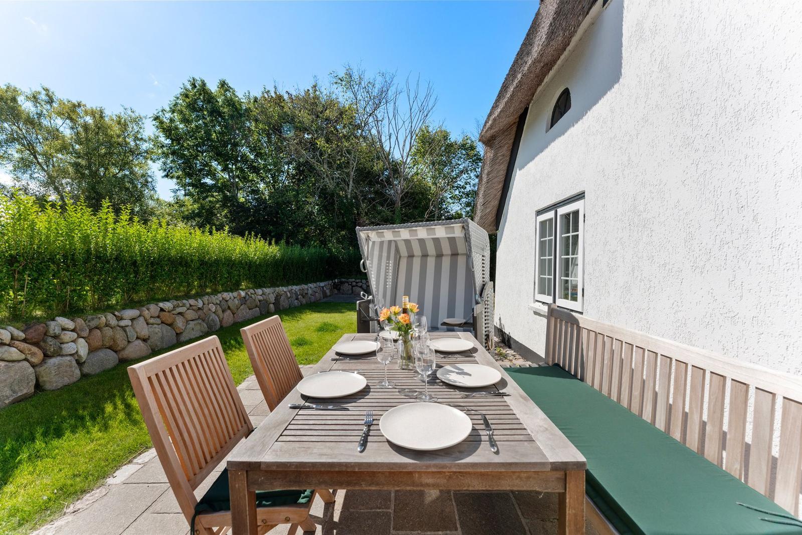 Gartenbereich mit gedecktem Tisch, Sitzbank und Pavillon neben weißer Hauswand.