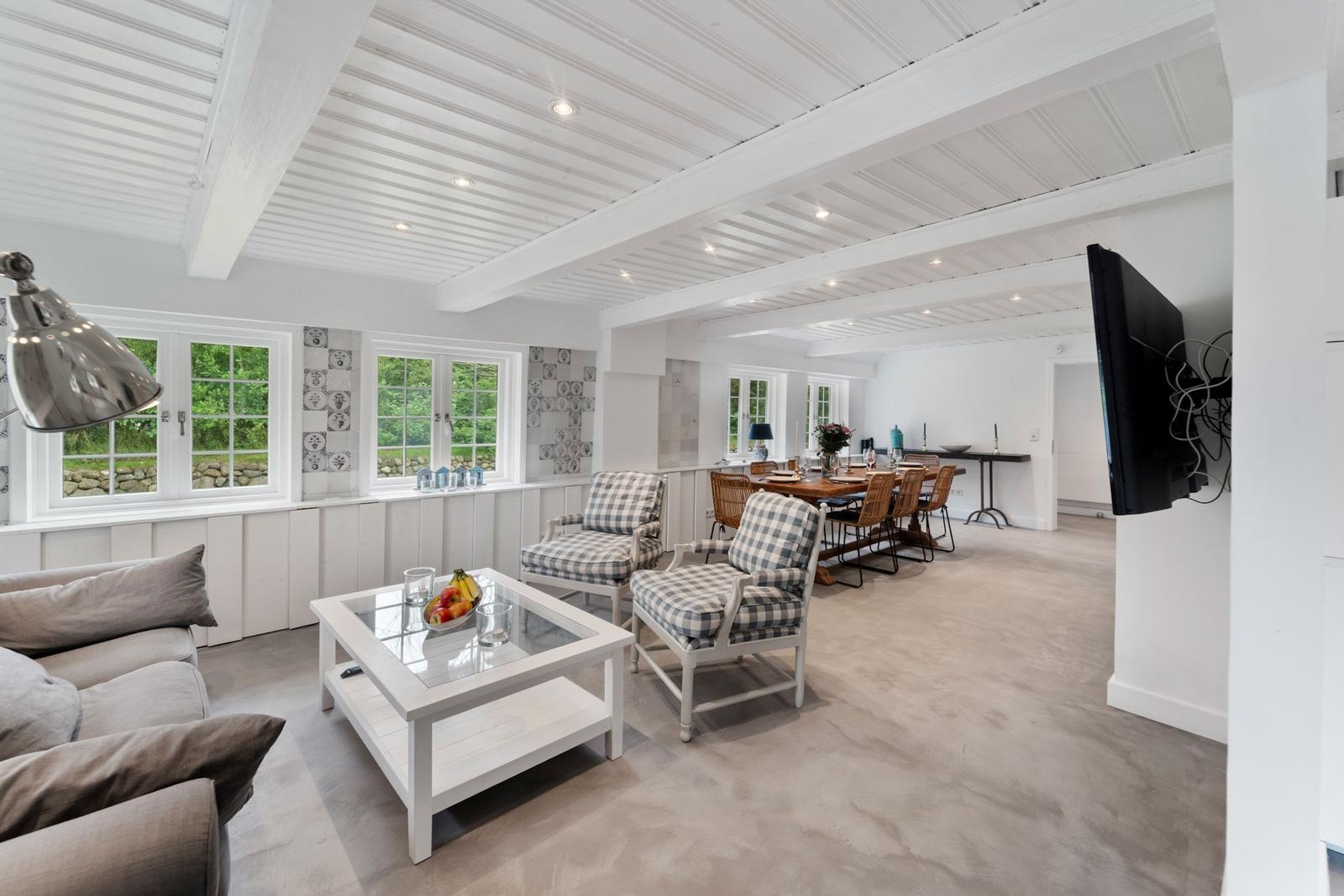 Wohn- und Essbereich mit weißen Wänden, Holzbalken und Fenstern mit Blick auf Grün.