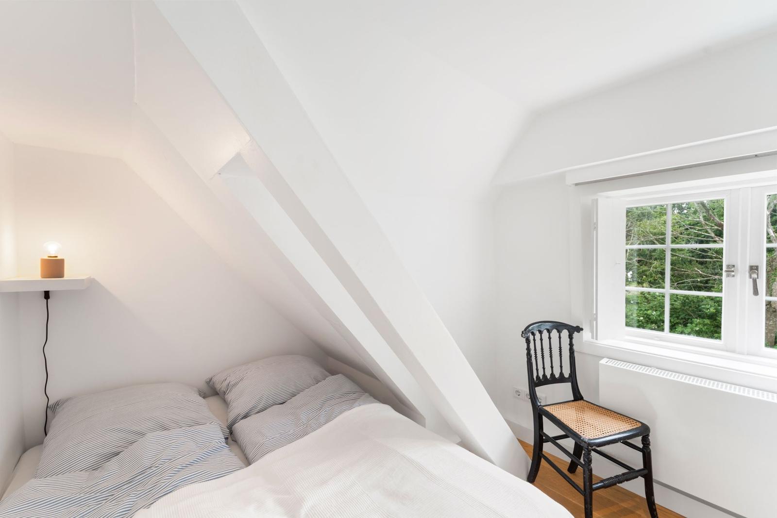 Schlafzimmer mit Dachschrägen, Bett, Stuhl und Fenster mit Blick auf Bäume.