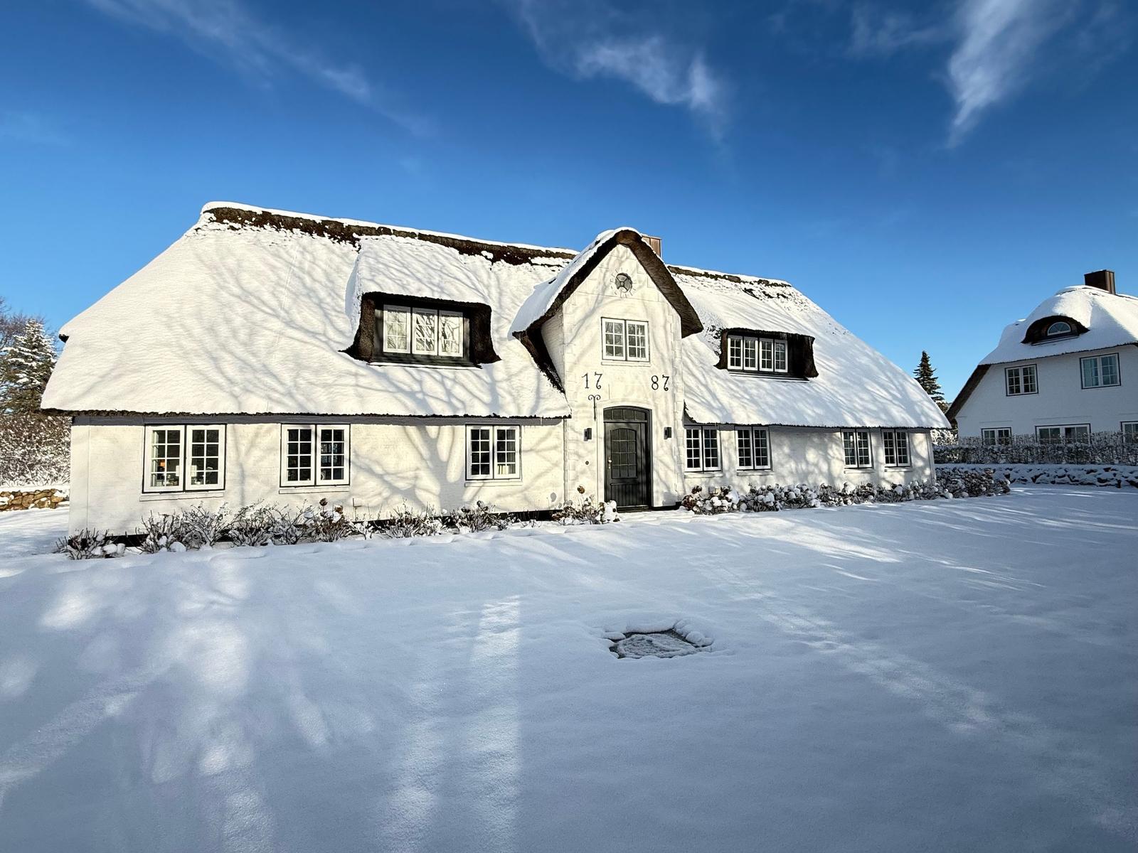 Schneebedecktes weißes Haus mit strohgedecktem Dach und mehreren Fenstern.
