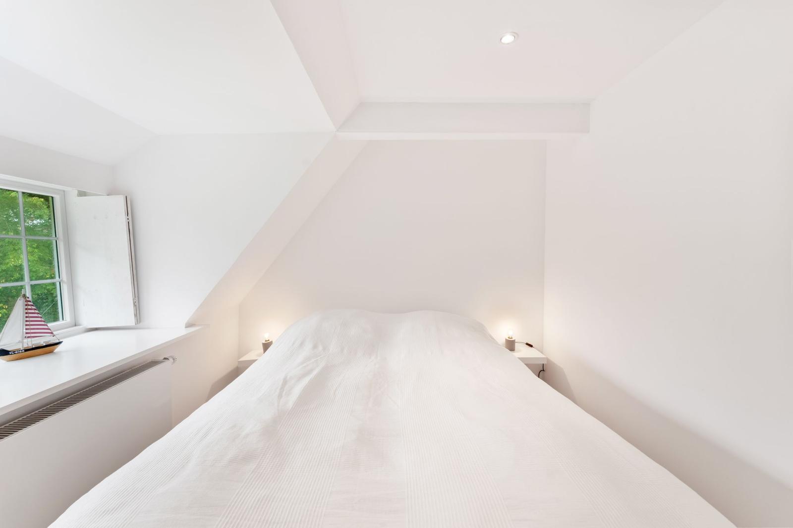 Schlafzimmer mit Dachschrägen, Bett und Fenster mit Segelbootmodell.