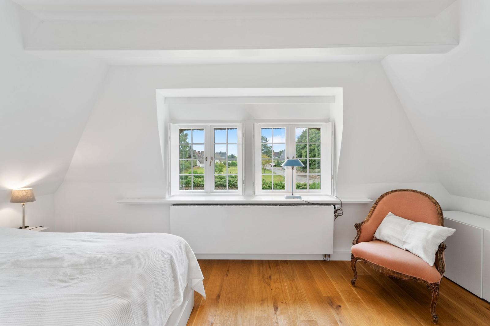 Schlafzimmer mit Dachschrägen, Fenster und Holzboden. Ein Bett, Stuhl und Lampen sind sichtbar.