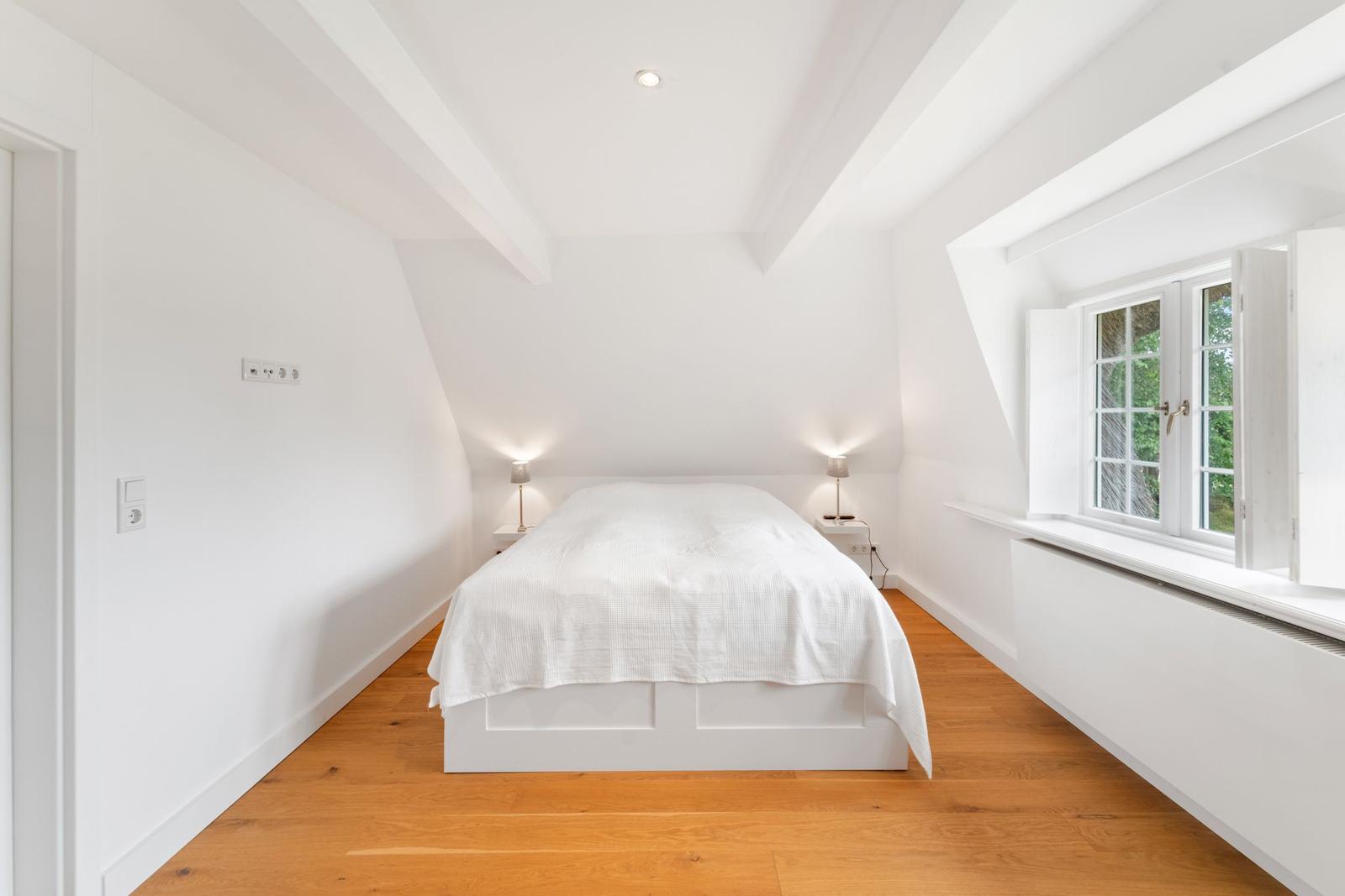Schlafzimmer mit Dachschrägen, weißem Bett und Holzboden. Fenster mit Blick auf Grün.