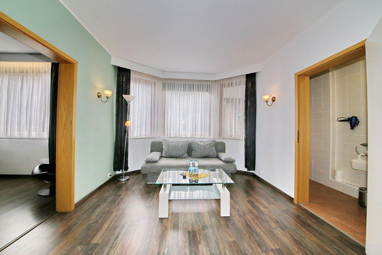 Wohnzimmer mit Sofa, Glas-Tisch und Blick auf Balkon. Nebenraum mit Dusche und Toilette.