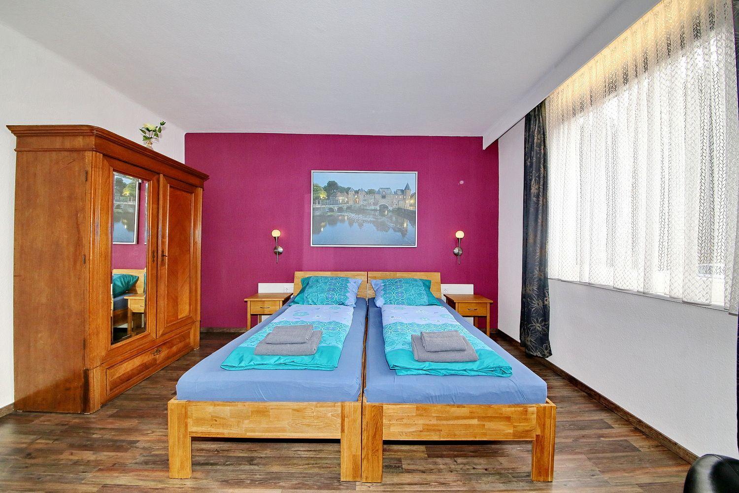 Zwei Betten mit blauen Laken stehen vor einer magentafarbenen Wand. Holzmöbel und Fenster mit Vorhängen.