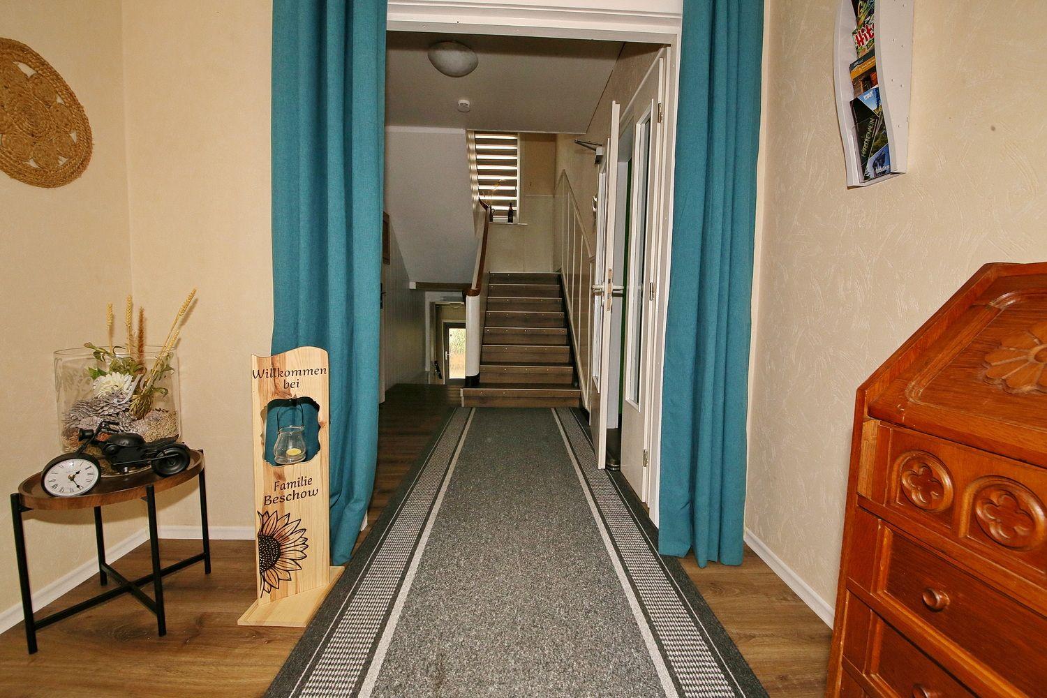 Eingangsbereich mit Teppich, Blauvorhängen und Holzschrank. Treppenhaus im Hintergrund.