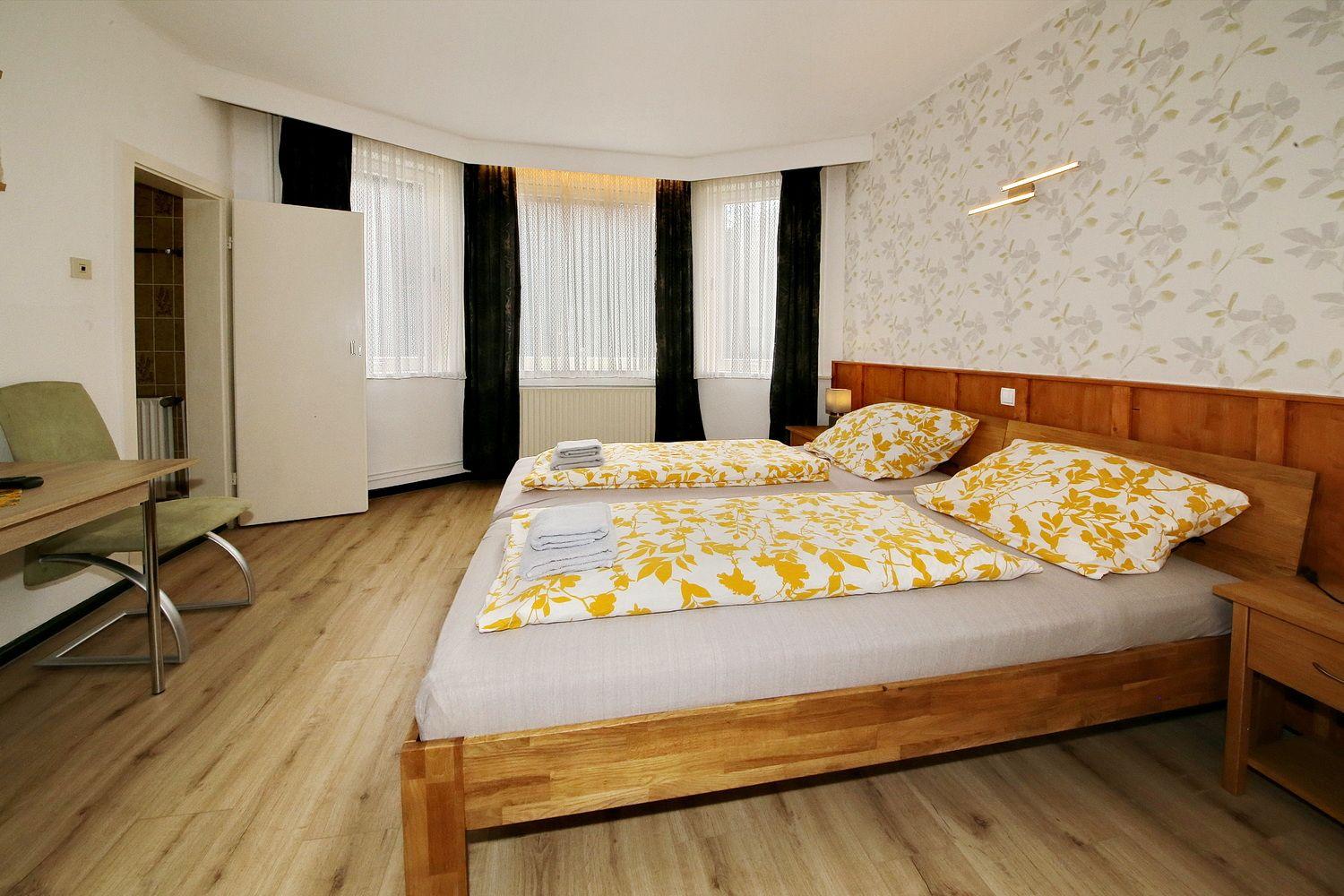 Zimmer mit Doppelbett, Holzboden und Fenstern mit Vorhängen.