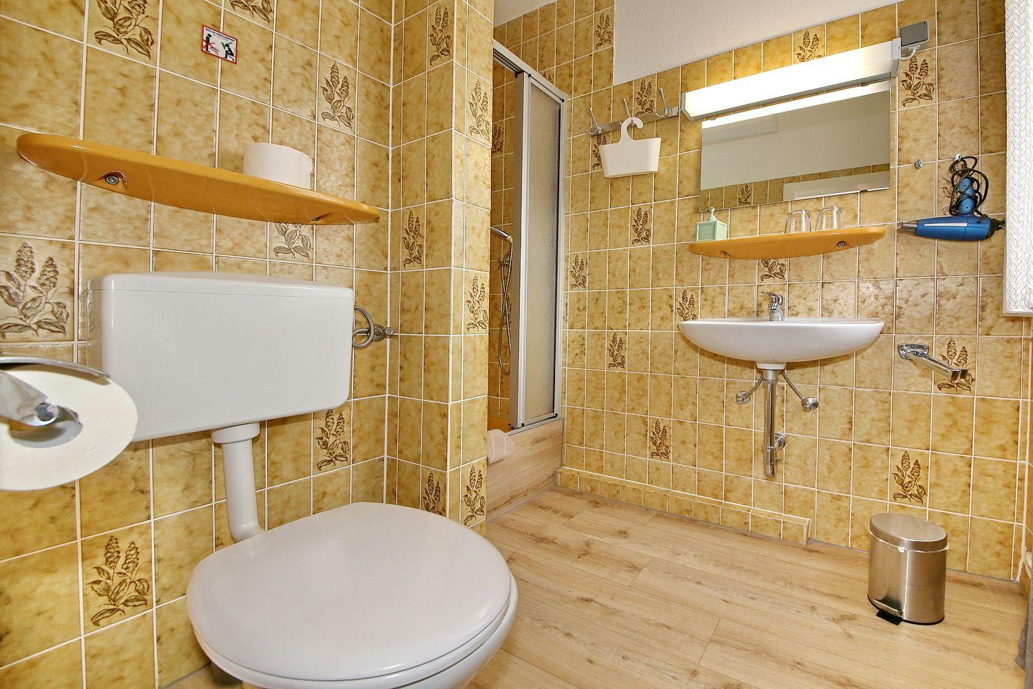 Badezimmer mit Toilette, Waschbecken und Dusche. Wandfliesen mit Pflanzenmotiv.