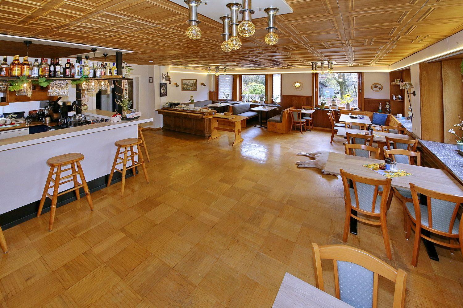 Großer Raum mit Holzboden, Tischen, Stühlen und Bar. Decke mit Holzverkleidung und Lampen.