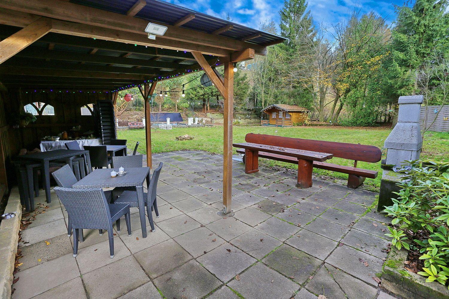 Terrasse mit Tischen, Stühlen und Bank unter Pavillon. Grünfläche und Wald im Hintergrund.