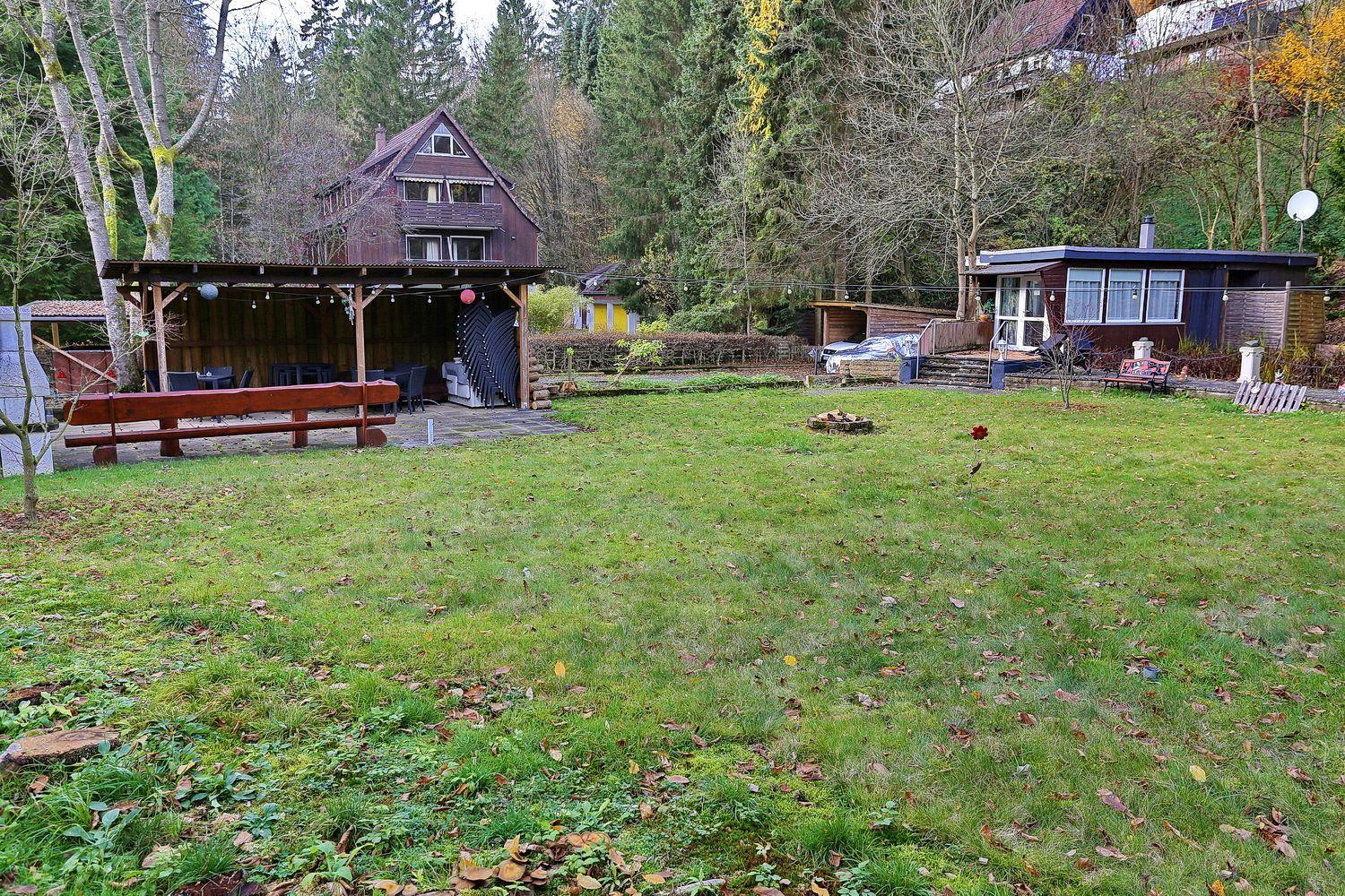 Grüner Garten mit Holzveranda, Terrasse und kleinem Haus im Hintergrund.