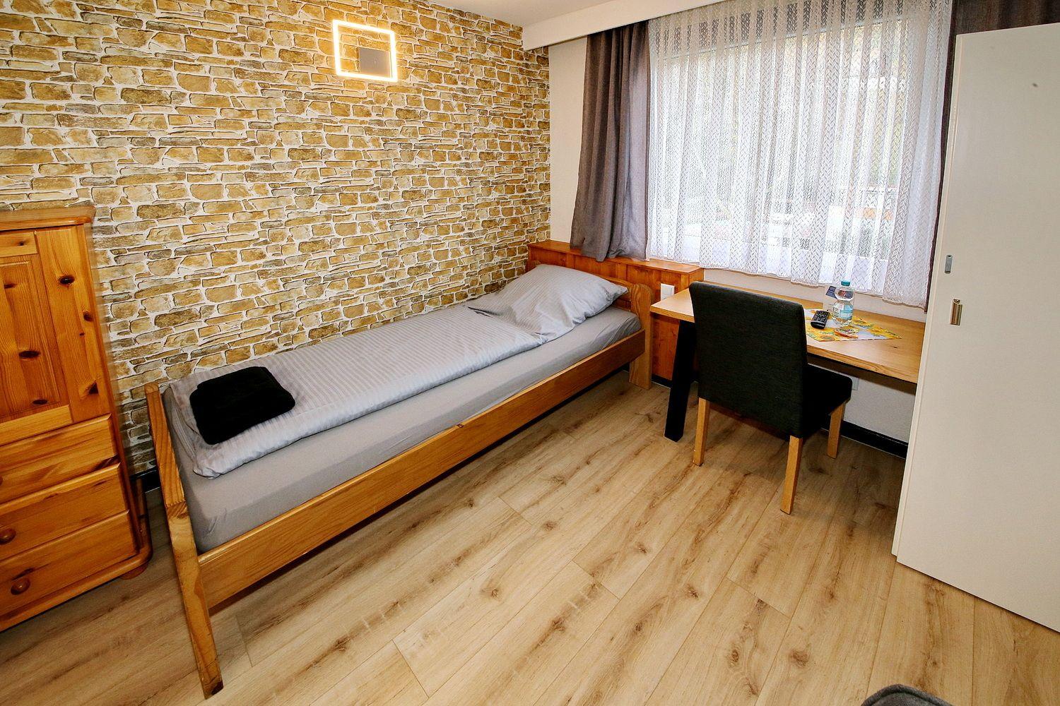 Ein Zimmer mit Holzbett, Schreibtisch und Steinwand. Fenster mit Vorhängen.