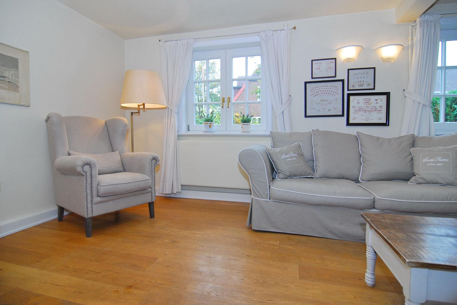 Wohnzimmer mit grauem Sofa, Sessel und Holzboden. Fenster mit weißen Vorhängen.