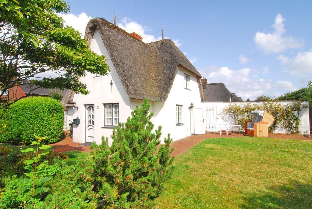 Weiße Hütte mit Strohdach, Garten und Terrasse mit Gartenmöbeln.