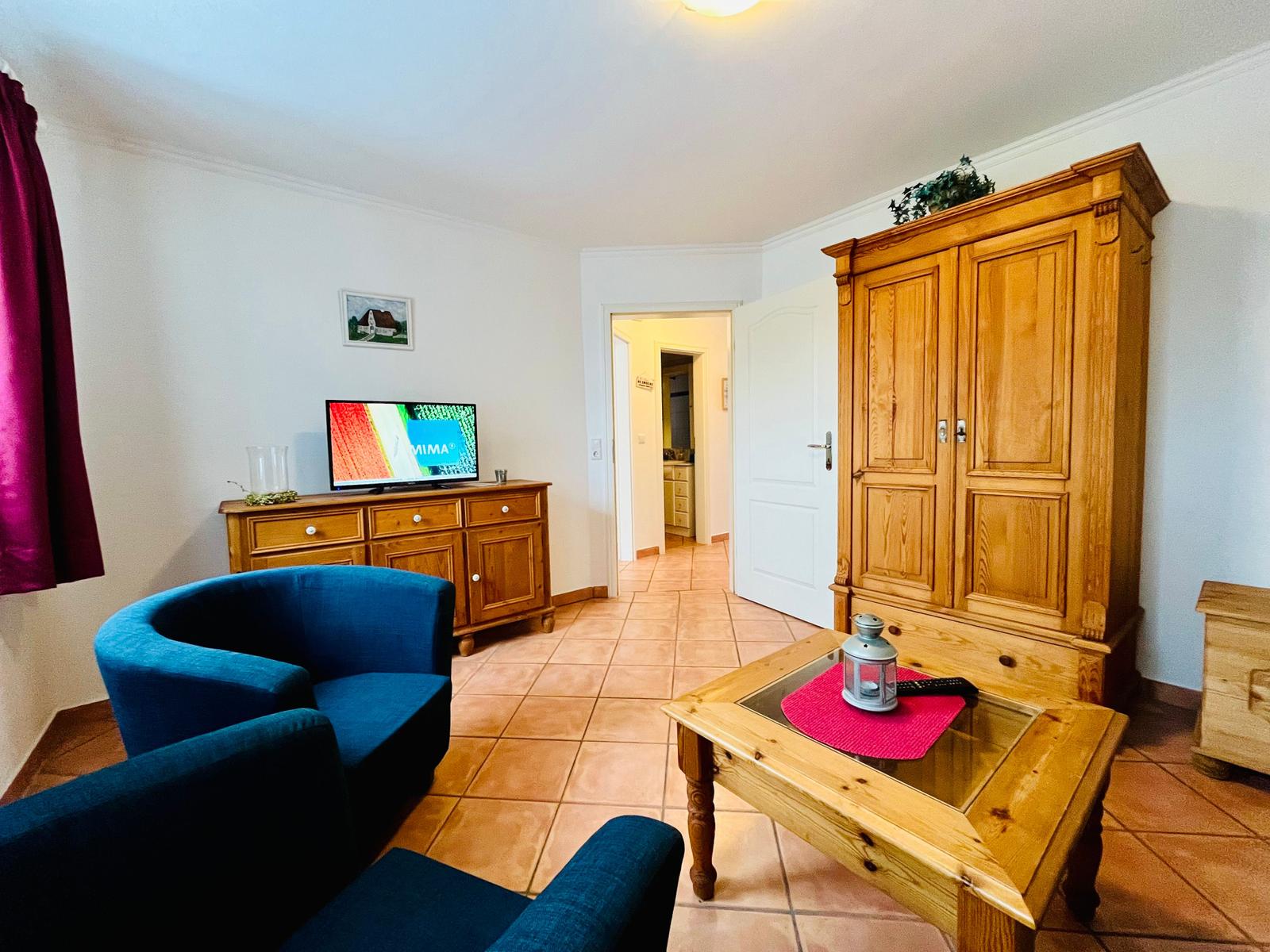 Wohnzimmer mit blauen Sesseln, Holzmöbeln und TV. Tür führt in die Küche.