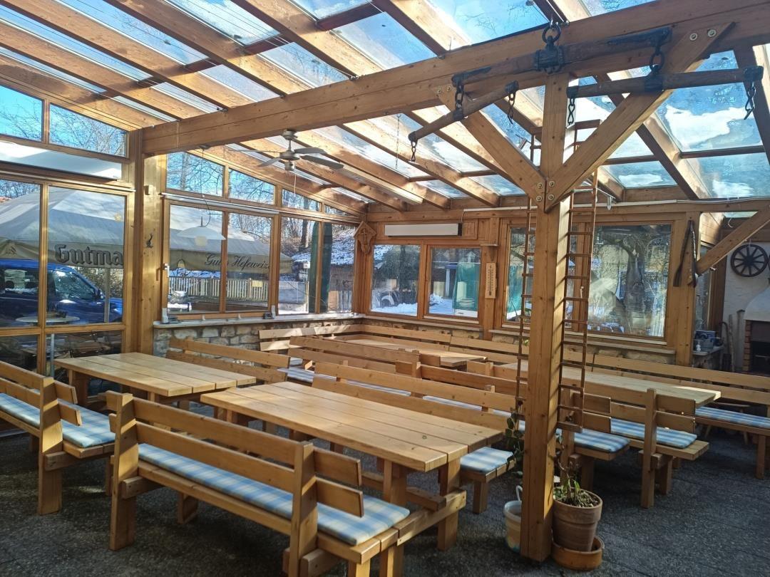 Holzterrasse mit Tischen und Bänken unter Glasdach. Außenbereich mit Blick auf Garten.