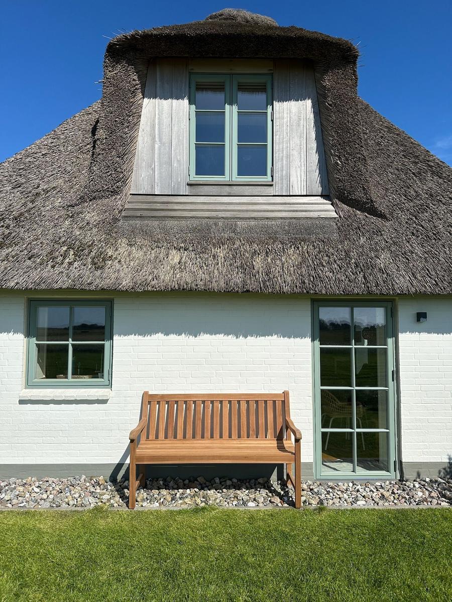 Haus mit Strohdach, Holzbank und grünen Fenstern vor weißem Mauerwerk.