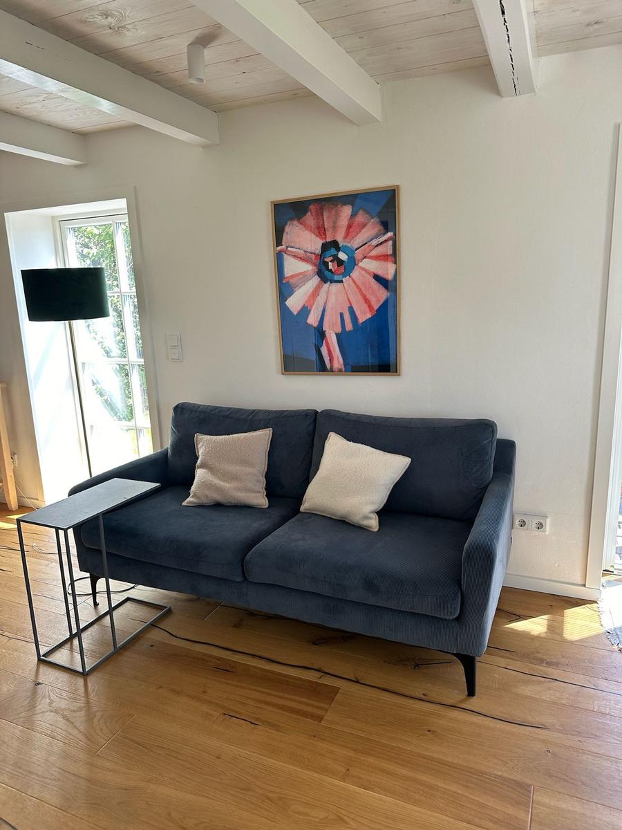 Ein dunkelblaues Sofa mit zwei Kissen steht auf Holzdielen vor einer Wand mit einem Bild.