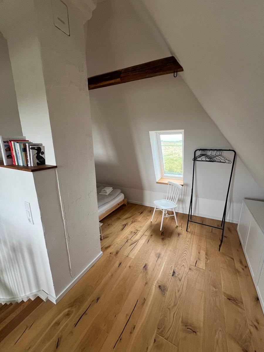 Zimmer mit Holzboden, Bett, Stuhl und Kleiderständer unter Dachschräge.