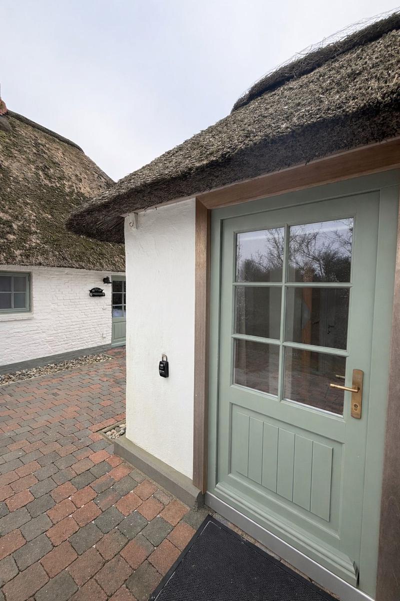 Haus mit Strohdach und grüner Tür. Vor dem Haus liegt ein gepflasterter Hof.