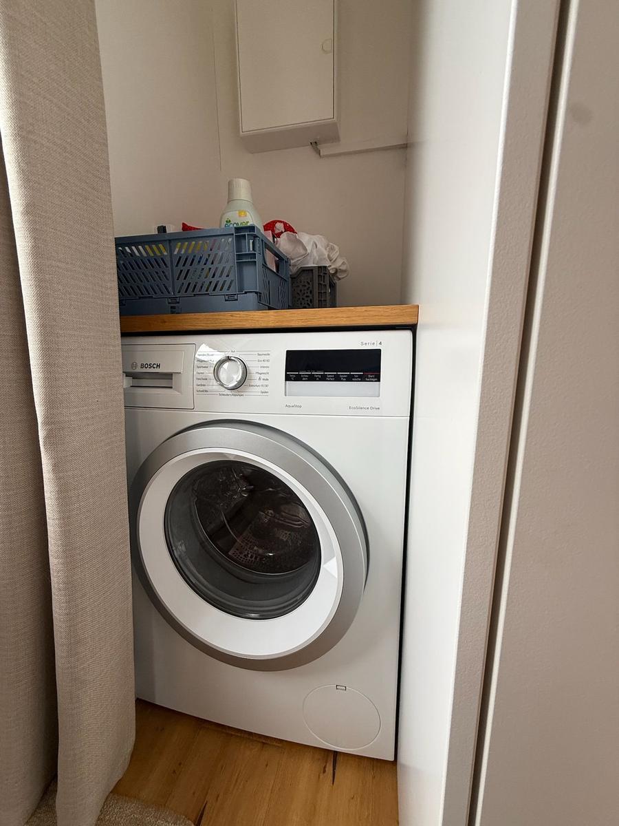 Waschmaschine in einem kleinen Raum mit Holzboden und Vorhang.