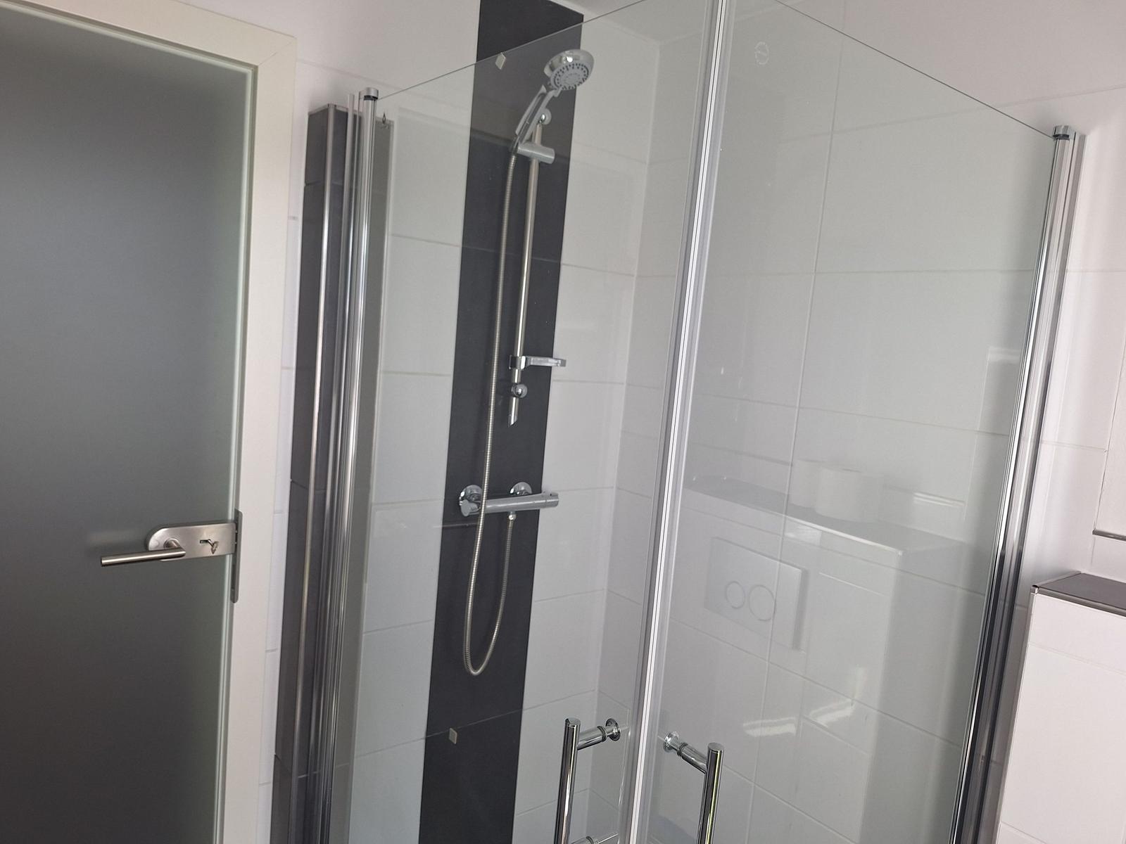 Shower with glass enclosure and handheld shower, white tiles and door on left