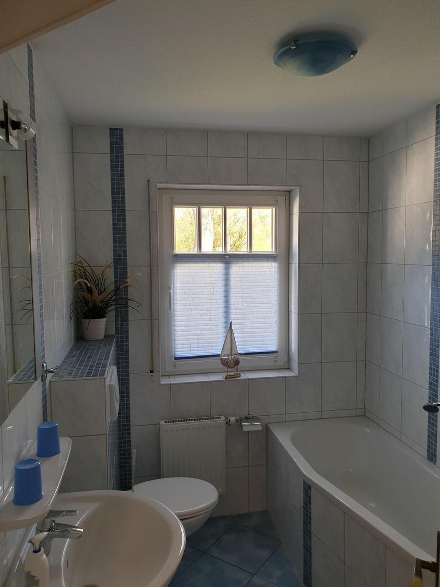 Badezimmer mit Badewanne, Toilette und Waschbecken. Fenster mit Jalousie und Segelboot-Dekoration.