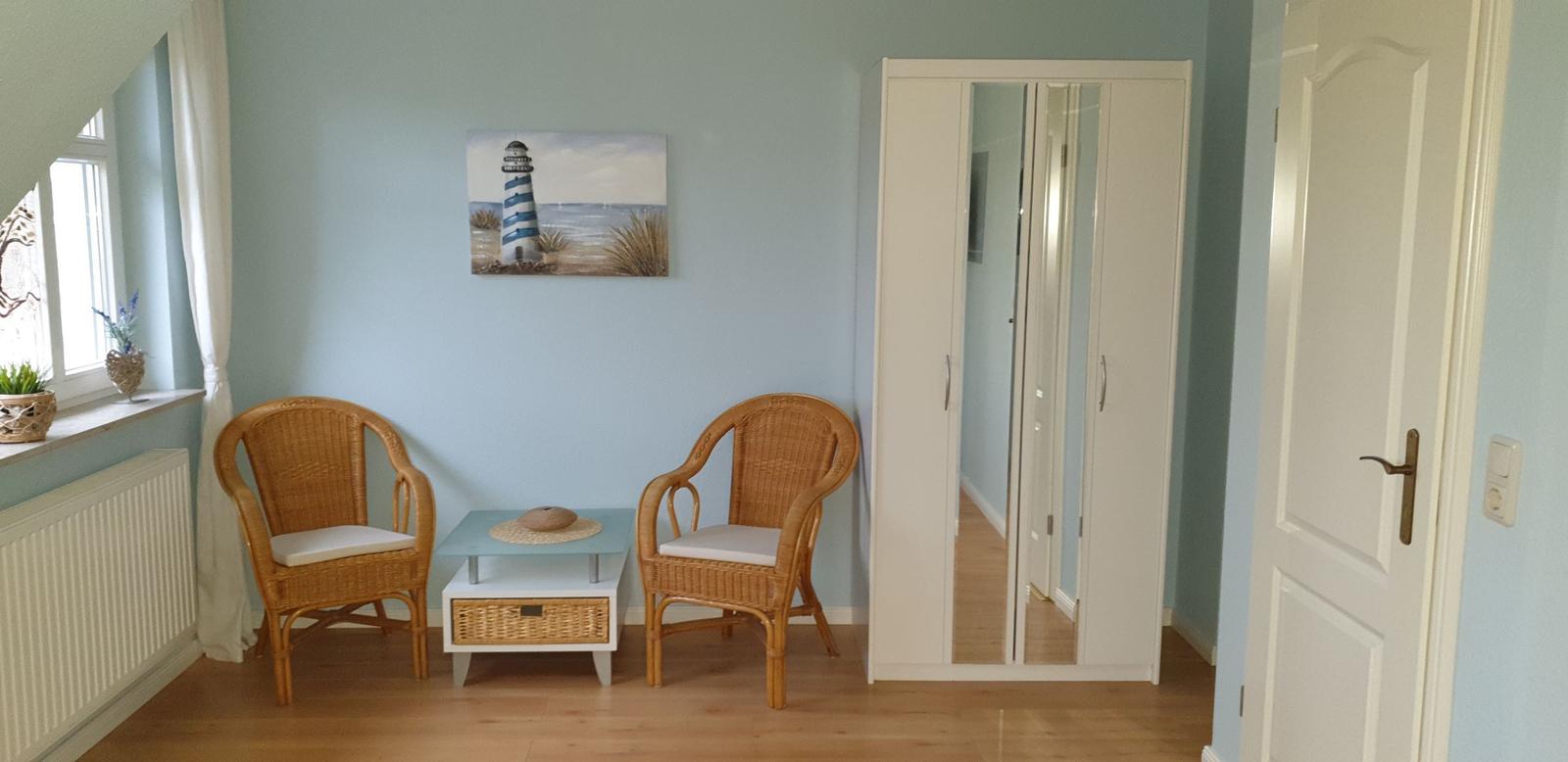 Zimmer mit zwei Rattanstühlen, Tisch und Kleiderschrank. Wandbild mit Leuchtturm.
