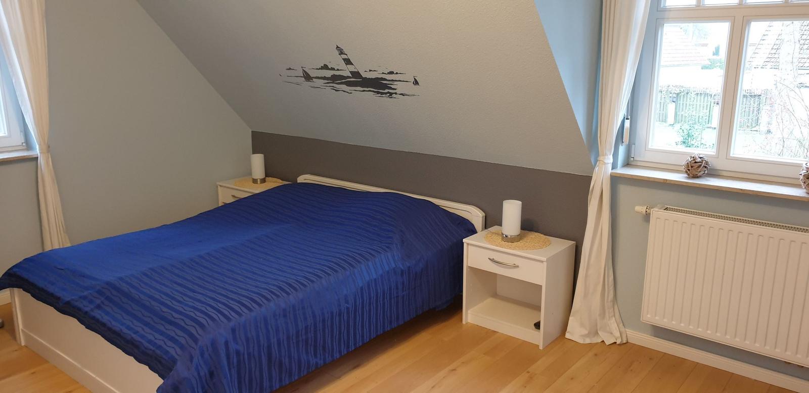 Schlafzimmer mit Dachschräge, Bett und Nachttisch. Wandbild zeigt Leuchtturm und Segelboote.