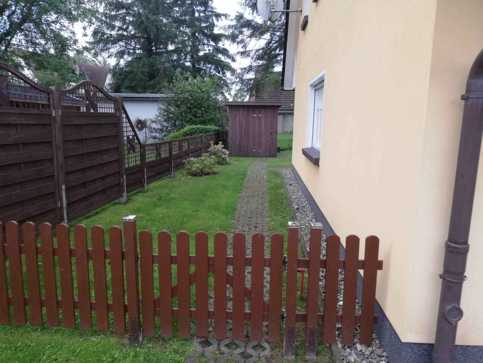 Hinterhof mit Rasen, Pflasterweg und Holzzaun. Hauswand und Garten sind sichtbar.