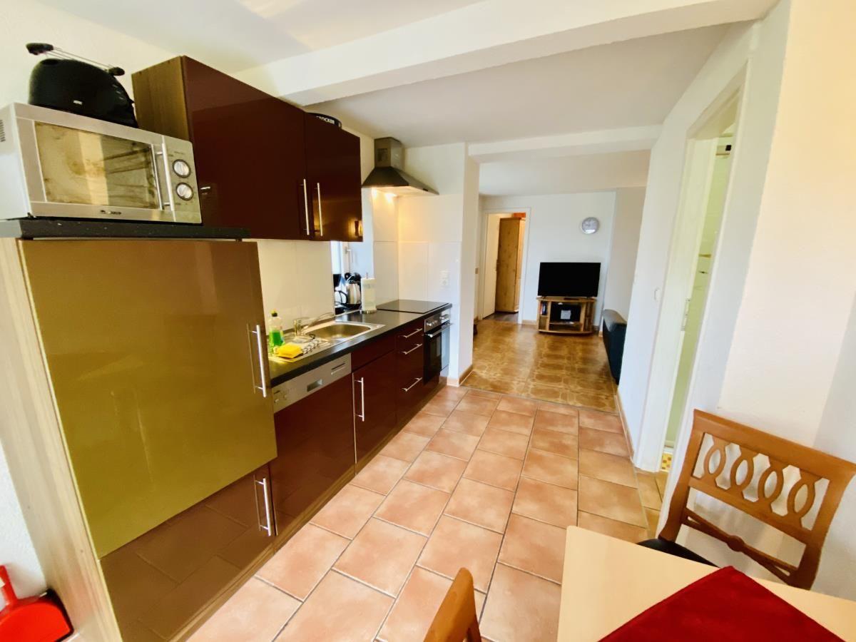 Küche mit dunklen Schränken und Terrakottaboden. Blick in Wohnzimmer mit Fernseher.