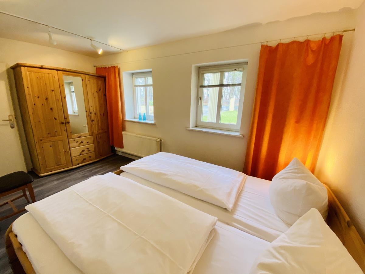 Zimmer mit zwei Betten, Holzschrank und Fenstern mit orangefarbenen Vorhängen.