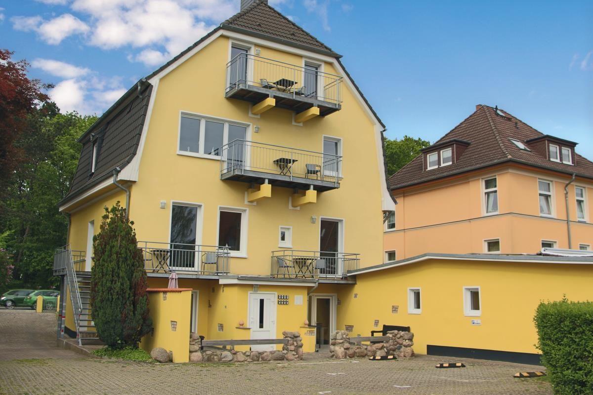 Gelbes Haus mit Balkonen und Terrassen. Umgeben von Bäumen und einem gepflasterten Hof.