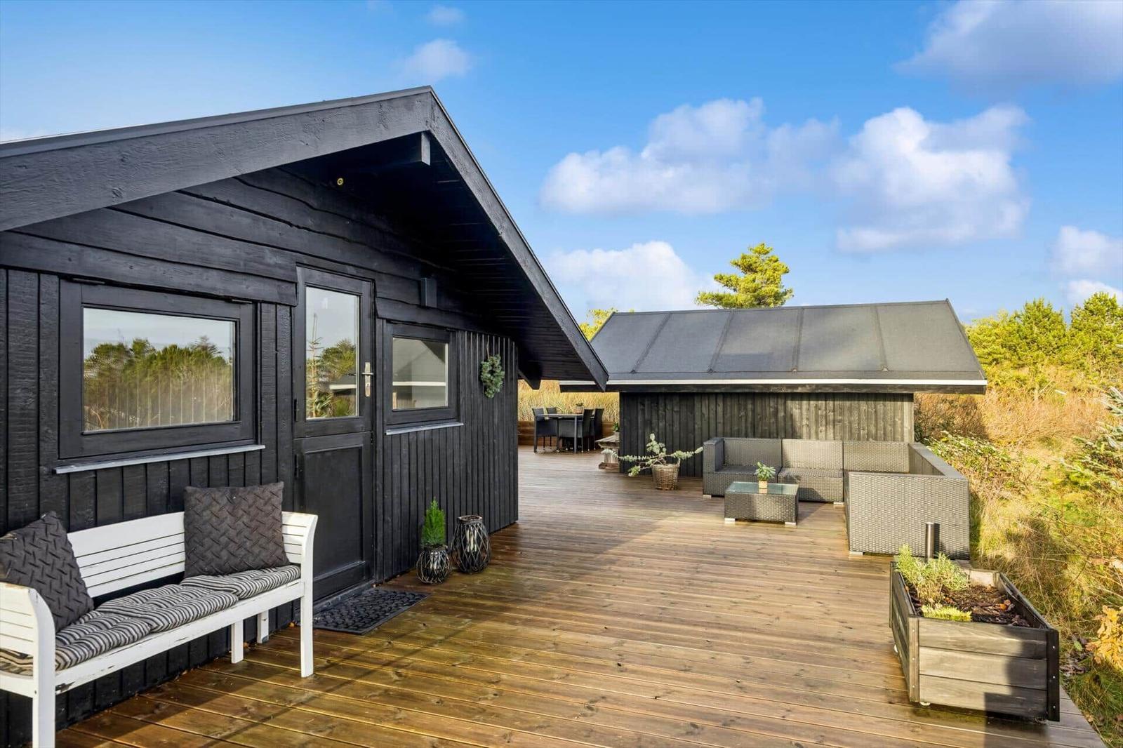 Black wooden cabin with large wooden terrace, garden furniture, and view of forest.